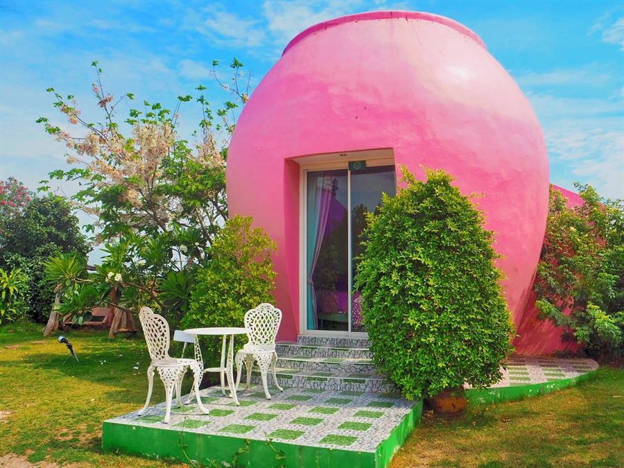 บ้านทรงโอ่งสีชมพู พร้อมโต๊ะเก้าอี้สีขาวหน้าบ้าน ท่ามกลางสวนและท้องฟ้าสดใส
