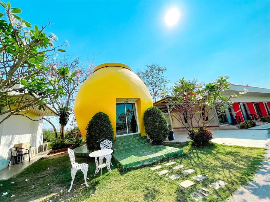 บ้านทรงโอ่งสีเหลืองสดใต้แสงแดด มีโต๊ะเก้าอี้เหล็กดัดสีขาวและสนามหญ้า
